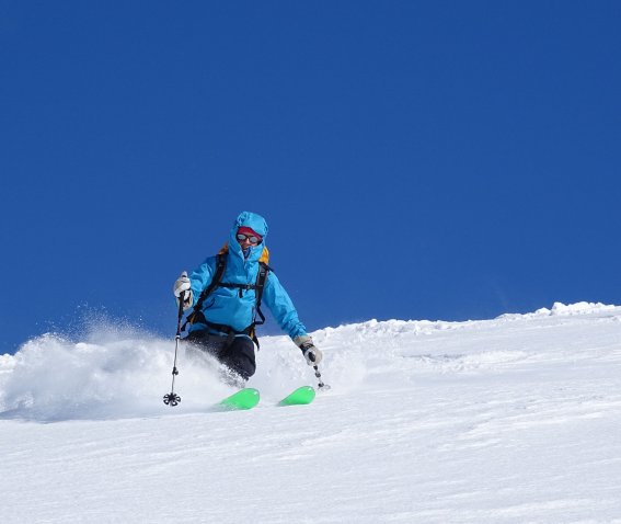 Skifahrer zieht kontrollierte Schwünge durch frischen Tiefschnee unter klarem, blauem Himmel.