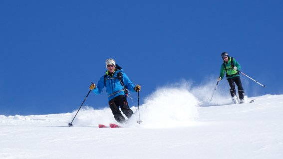 Zwei Skifahrer ziehen dynamische Spuren durch frischen Pulverschnee unter klarem, tiefblauem Himmel.