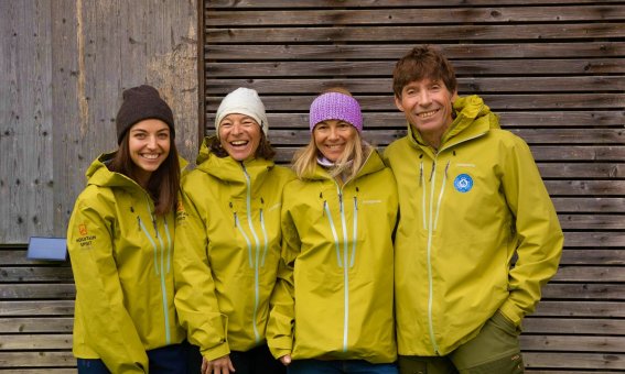 Mountain-Spirit-Team in grünen Outdoorjacken steht lachend vor Holzwand – Bergführer und Organisationsteam für Ski- und Abenteuerreisen.