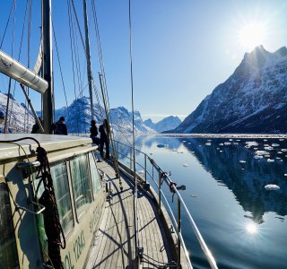 Leute stehen auf einem Boot und schauen auf die Berge. Die Sonne scheint auf das Wasser und die Eisstücke.