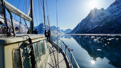 Leute stehen auf einem Boot und schauen auf die Berge. Die Sonne scheint auf das Wasser und die Eisstücke.