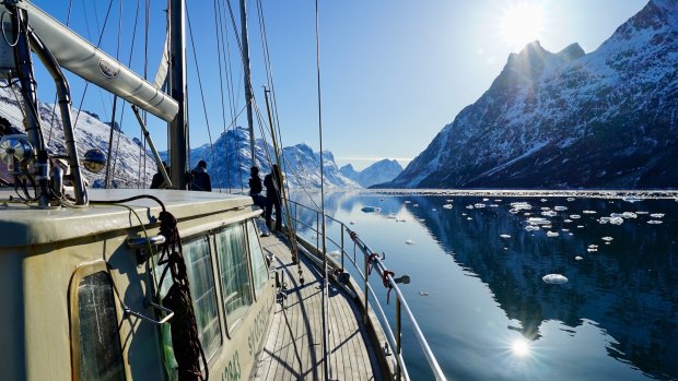 Schifffahrt im Fjord während des Tages Leute stehen auf einem Boot und schauen auf die Berge. Die Sonne scheint auf das Wasser und die Eisstücke.