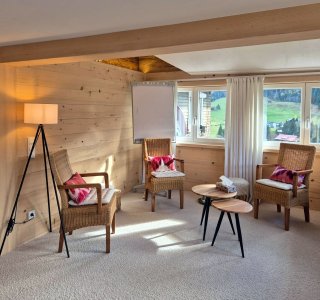 Gemütliches Wohnzimmer mit Holzwänden, Blick auf die Berge und sonnigem Licht, ideal für Entspannung und gesellige Stunden.