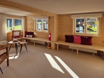 Diese gemütliche Stube hat große Fenster, helles Holz und wartet auf entspannte Stunden. Der Ausblick ins Grüne ist einladend.