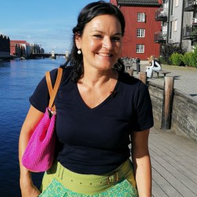 Eine freundliche Frau geht am Wasser entlang und genießt das sonnige Wetter. Im Hintergrund sind Häuser zu sehen.