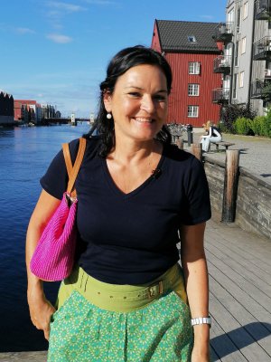 Eine freundliche Frau geht am Wasser entlang und genießt das sonnige Wetter. Im Hintergrund sind Häuser zu sehen.