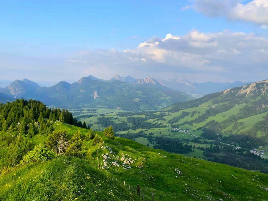 Die Landschaft zeigt grüne Hügel und Berge unter blauem Himmel. Wolken ziehen über die Gipfel. Es ist ein sonniger Sommertag.