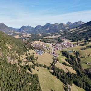 Das Bild zeigt eine weite Berglandschaft mit einem kleinen Dorf in den Alpen. Es ist tagsüber mit klarem Himmel.