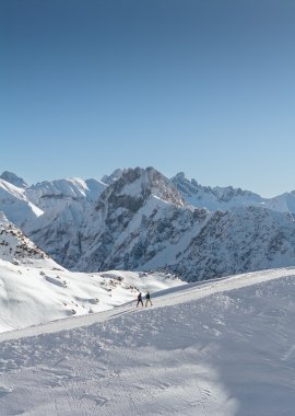 Zwei kleine Personen wandern auf dem verschneiten Zeigersattel am Nebelhorn. Im Hintergrund sieht man die imposante Höfats.