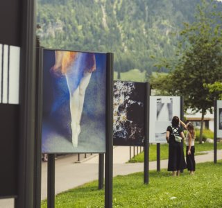 Besucher schauen sich Kunstwerke im Freien an. Die Ausstellung zeigt verschiedene Bilder vor einer Bergkulisse.