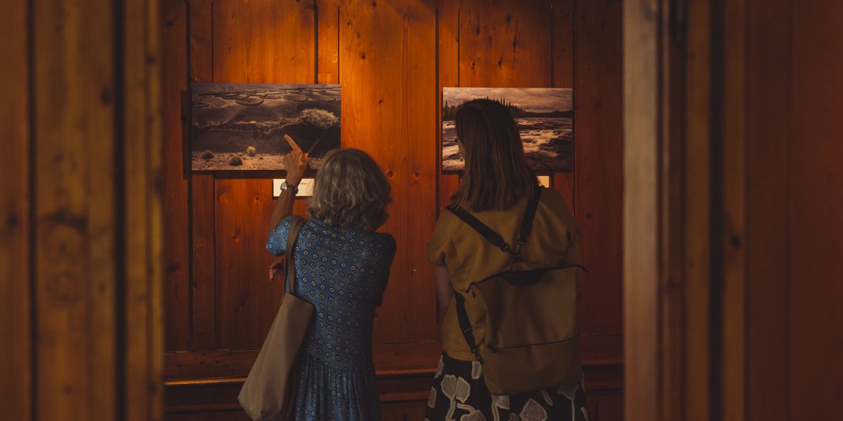 Zwei Frauen schauen sich Kunstwerke in einer Galerie an. Die Wände sind aus Holz und die Beleuchtung ist sanft.