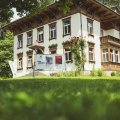 Das Haus steht in einer Gartenlandschaft. Es gibt Fenster mit Blumen und ein Schild vor dem Gebäude.