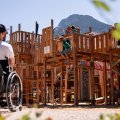 Kinder spielen auf der großen Spielburg des inklusiven Spielplatzes am Fuggerpark. Im Vordergrund ein Rollstuhlfahrer.