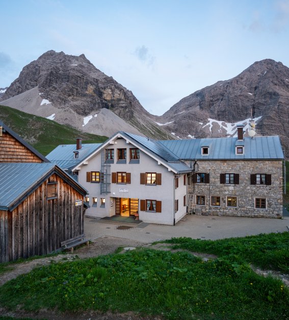 Rappenseehütte Ein Bauernhof steht in den Alpen. Die Berge sind sichtbar. Es wird dunkel und die Fenster leuchten.