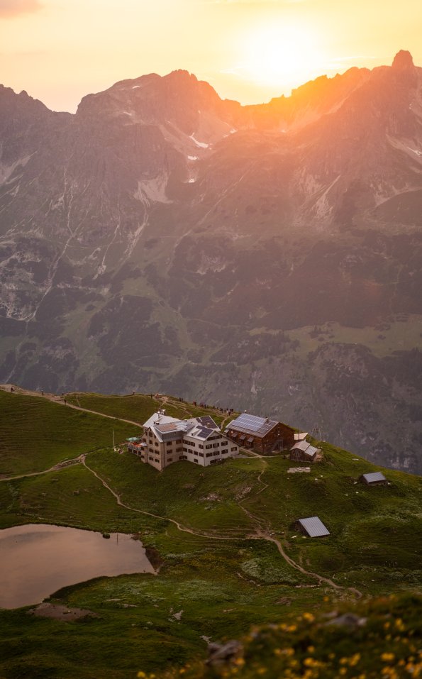 Die Berghütte liegt auf einer Wiese und wird von Bergen umgeben. Die Sonne geht unter und färbt den Himmel orange.