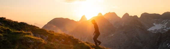 Eine Person wandert auf einem Hügel während des Sonnenuntergangs. Die Berge sind im Hintergrund sichtbar.
