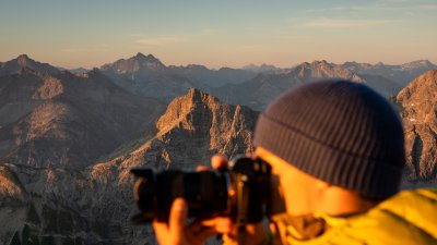 Ein Mensch steht auf einem Berg und fotografiert die Landschaft in den Alpen. Es ist Abend und die Sonne scheint.