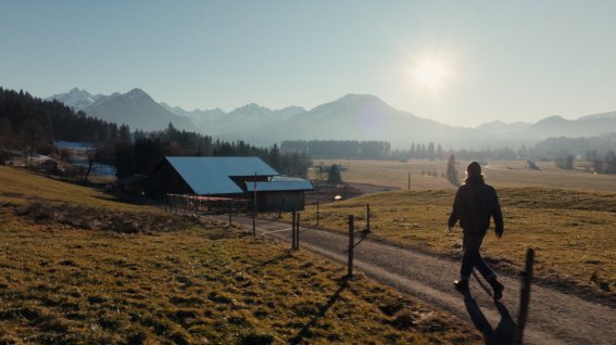 Ein Mann geht auf einem Weg durch eine offene Landschaft. Die Sonne geht im Hintergrund auf und beleuchtet die Berge.