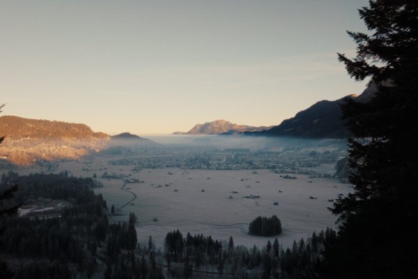 Ein Tal mit Bäumen und Schnee ist am Morgen zu sehen. Die Sonne scheint über die Berge im Hintergrund.