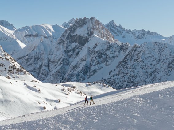Zwei kleine Personen wandern auf dem verschneiten Zeigersattel am Nebelhorn. Im Hintergrund sieht man die imposante Höfats.