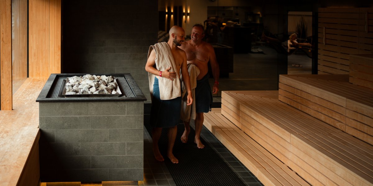 Zwei Männer gehen in eine Sauna mit Holzbänken und Steinen. Es ist ein entspannender Moment in einem Wellnessbereich.