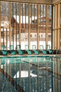 Ein Schwimmbad mit blauen Liegen hat große Fenster. Draußen ist eine verschneite Landschaft mit Häusern.