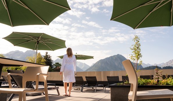 Eine Frau steht auf einer Terrasse in den Bergen und schaut in die Ferne. Die Sonne scheint und es gibt Sonnenschirme.