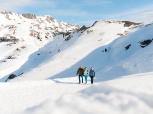 Drei Personen gehen auf einem Pfad in den verschneiten Alpen. Der Himmel ist klar und die Sonne scheint.