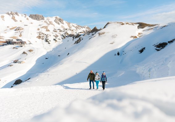 Winterwandern auf dem Zeigersattel Drei Personen gehen auf einem Pfad in den verschneiten Alpen. Der Himmel ist klar und die Sonne scheint.