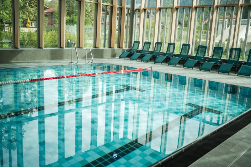 Im Schwimmbad gibt es Wasser, Liegen und große Fenster. Es sieht hell und einladend aus.