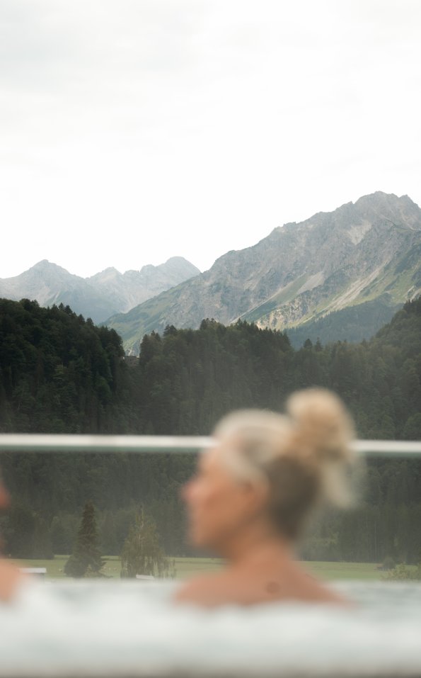 Zwei Personen sitzen im Wasser und schauen auf die Berge. Es ist eine ruhige Landschaft mit vielen Bäumen.
