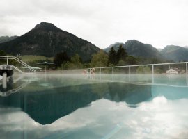 Im Hintergrund sind Berge zu sehen, während Dampf über einem Schwimmbad aufsteigt. Das Wetter wirkt bewölkt und kühl.