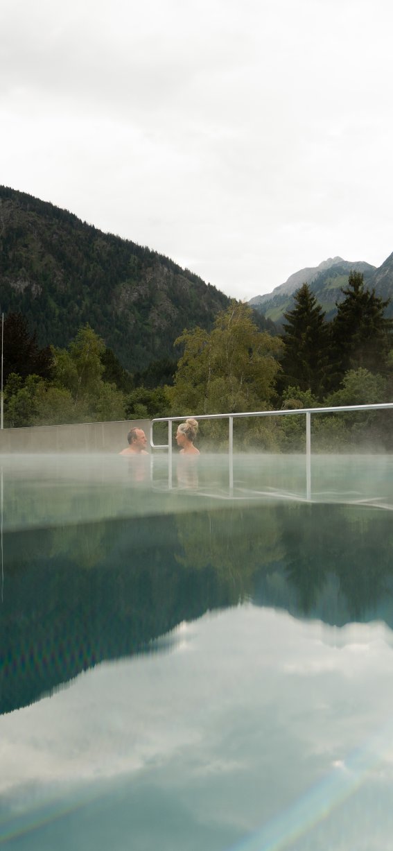 Im Hintergrund sind Berge zu sehen, während Dampf über einem Schwimmbad aufsteigt. Das Wetter wirkt bewölkt und kühl.