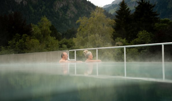 Drei Personen genießen das warme Wasser in einem Außenpool in den Bergen an einem bewölkten Tag.