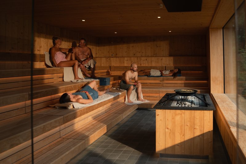 Menschen sitzen und liegen in einer Sauna mit Holzbank und Kamin. Es ist warm und gemütlich.