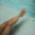 Eine Person sitzt am Beckenrand und hält die Füße im Wasser, während es regnet. Es gibt Wassertropfen auf der Oberfläche.