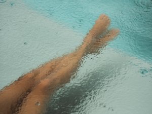 Eine Person sitzt am Beckenrand und hält die Füße im Wasser, während es regnet. Es gibt Wassertropfen auf der Oberfläche.