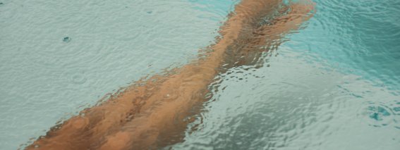 Eine Person sitzt am Beckenrand und hält die Füße im Wasser, während es regnet. Es gibt Wassertropfen auf der Oberfläche.