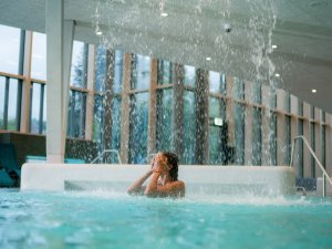 Eine Person steht unter einem Wasserfall in einem Innenpool. Der Ort hat große Fenster und viel Tageslicht.