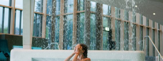 Eine Person steht unter einem Wasserfall in einem Innenpool. Der Ort hat große Fenster und viel Tageslicht.