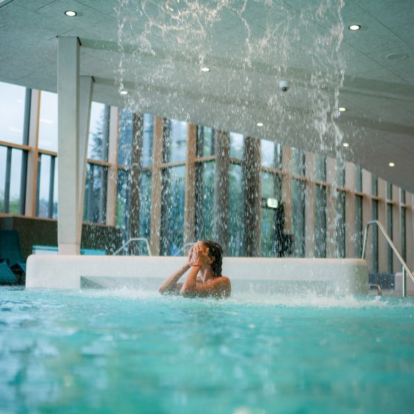 Eine Person steht unter einem Wasserfall in einem Innenpool. Der Ort hat große Fenster und viel Tageslicht.