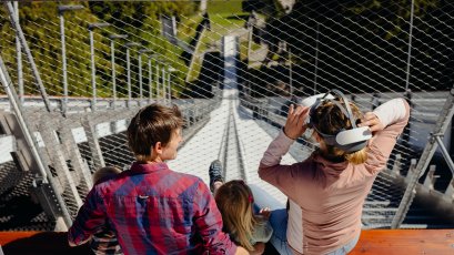 Familie auf dem Absprungbalken Eine Familie sitzt zusammen auf einer Bank am Aussichtspunkt und blickt in die Ferne. Sie nutzen eine VR-Brille.
