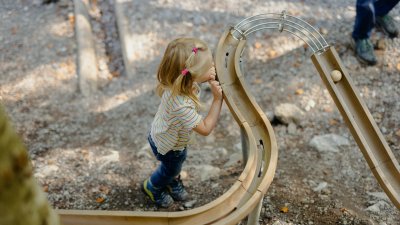 Ein Kind spielt mit einer kleinen Kugel auf einer Holzbahn im Freien. Es gibt Bäume und Erde um die Bahn herum.