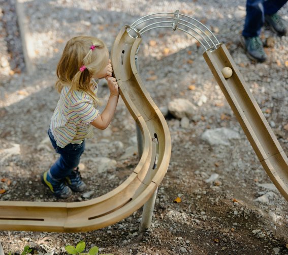 Kind spielt mit der Kugelbahn Ein Kind spielt mit einer kleinen Kugel auf einer Holzbahn im Freien. Es gibt Bäume und Erde um die Bahn herum.