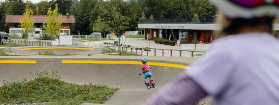 Ein Kind fährt mit einem Roller auf einer Strecke im Park. Im Hintergrund sind weitere Kinder und Gebäude.