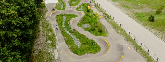 Der Skatepark befindet sich umgeben von Bäumen und Bergen. Einige Sportler nutzen die Anlagen zum Skaten und Fahren.