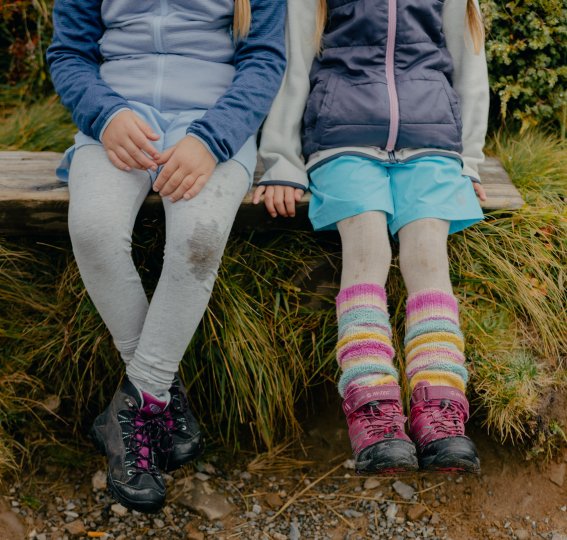 Zwei Kinder sitzen auf einer Bank in der Natur. Sie tragen bequeme Kleidung und Stiefel.