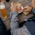Ein Mann sitzt an einem Holztisch und isst ein Gericht mit Brot und trinkt ein Bier. Es ist eine gesellige Atmosphäre.