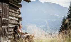 Ein Paar, das sich vor einer kleinen, alten Holzhütte sonnt. Sie genießen den Blick Richtung Berge sowie das Gefühl der Wärme, die die Sommersonne auf ihren Gesichtern hinterlässt. Im Vordergrund steht hohes Gras.