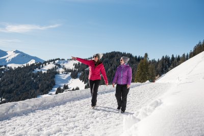 Zwei Frauen wandern bei Sonnenschein auf dem verschneiten Höhenpanoramaweg.